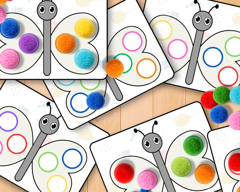 May include: A collection of butterfly-shaped educational cards with colorful pom-poms. Each card features a butterfly design with circles for pom-pom placement. The pom-poms are in various colors, including blue, purple, orange, and yellow.