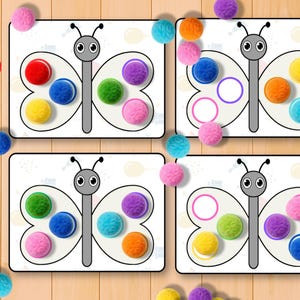 May include: Four white butterfly-shaped cards with colorful pom-poms and matching circles. The pom-poms are red, blue, yellow, green, purple, orange, and pink. The cards are on a light brown wooden surface.