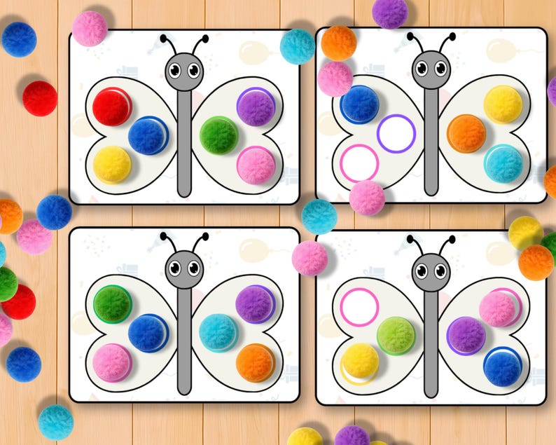 May include: Four white butterfly-shaped cards with colorful pom-poms and matching circles. The pom-poms are red, blue, yellow, green, purple, orange, and pink. The cards are on a light brown wooden surface.