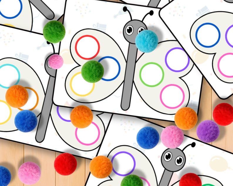 May include: Butterfly-shaped educational cards with colorful circles and matching pom-poms. The cards feature a gray butterfly outline with circles in red, orange, yellow, green, blue, and purple. Various colored pom-poms are scattered around the cards.