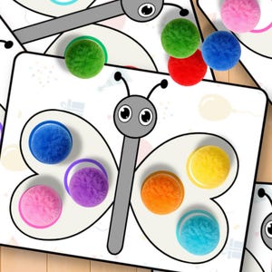 May include: Butterfly-shaped educational game with colorful pom-poms. The game features a white background with a butterfly outline and matching colored circles. Various colored pom-poms are scattered around the game, including green, red, blue, and pink.