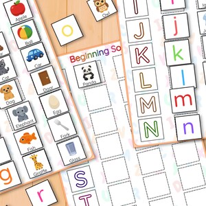May include: Educational alphabet flashcards with colourful illustrations. Each card features a letter and a corresponding image, such as an apple, ball, or car. The cards are designed for early learning and are arranged on a wooden surface.