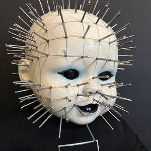 May include: A doll head with pale white skin and black painted eyes and lips, featuring numerous silver metal nails protruding from the surface in a grid pattern. The doll wears a black garment with a rectangular silver metal frame at the neck.