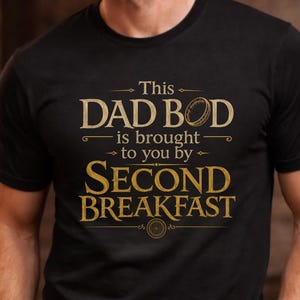 May include: Black t-shirt with gold text reading "This Dad Bod is brought to you by Second Breakfast." The shirt has a crew neck and short sleeves. The text is in a serif font.