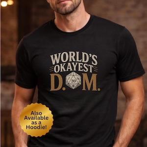 May include: Black t-shirt with the text "WORLD'S OKAYEST D.M." in a distressed, vintage style. The design includes a 20-sided die graphic. A gold sticker at the bottom says "Also Available as a Hoodie!"