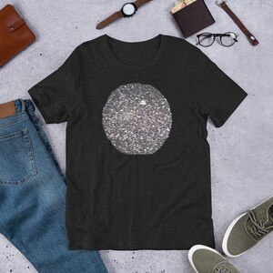 Puede incluir: Camiseta gris oscuro con un diseño de círculo plateado grande y brillante. La camiseta está hecha de un material suave y tiene mangas cortas. La camiseta se muestra sobre una superficie gris con otros accesorios.