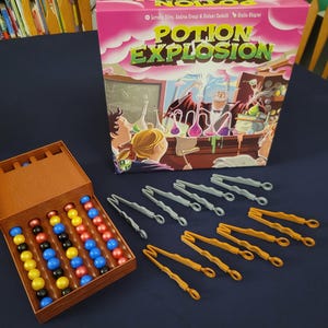 May include: The board game "Potion Explosion" is displayed, featuring a pink box with the game title and illustrations. A wooden-colored container holds colorful marbles. Gray and orange plastic tongs are also visible, suggesting a game of strategy and skill.
