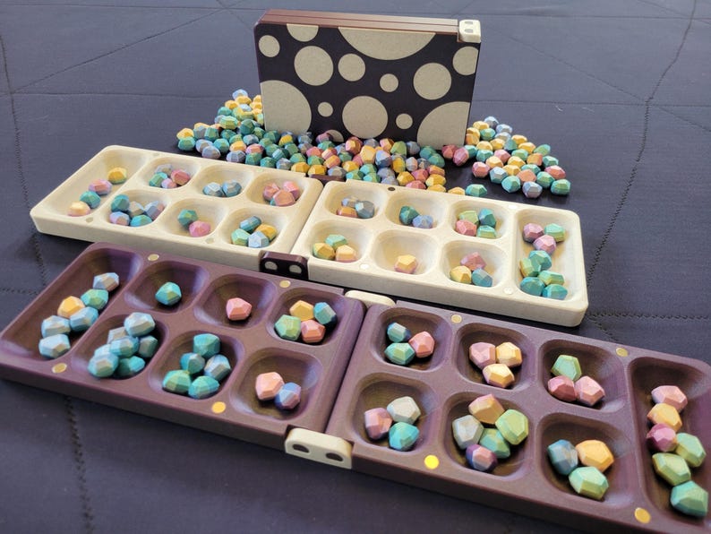 May include: A Mancala board game set with a purple and cream-coloured board, and a box with a polka dot design. The game pieces are small, multi-coloured stones. The game is set on a dark blue surface.