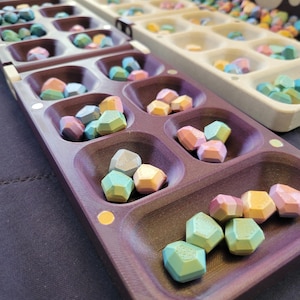 May include: Two Mancala game boards are shown, one purple and one beige, filled with multi-coloured, geometric stones. The stones are in shades of blue, green, pink, and yellow. The game is set on a dark surface.