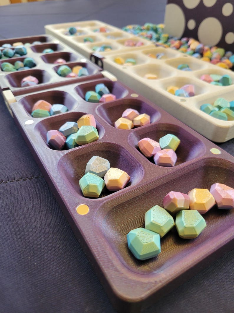 May include: Two Mancala game boards are shown, one purple and one beige, filled with multi-coloured, geometric stones. The stones are in shades of blue, green, pink, and yellow. The game is set on a dark surface.