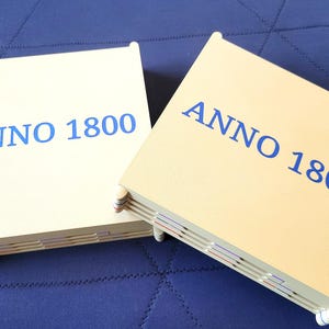 May include: Two cream-colored game boxes with the text "ANNO 1800" in blue. The boxes are rectangular with rounded corners and visible binding. The boxes are on a blue surface.