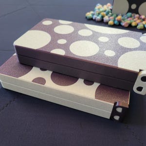 May include: Two rectangular boxes with a polka dot pattern in white and dark brown. The boxes are stacked on top of each other. The edges of the boxes are dark brown and white. The boxes are on a dark blue surface.