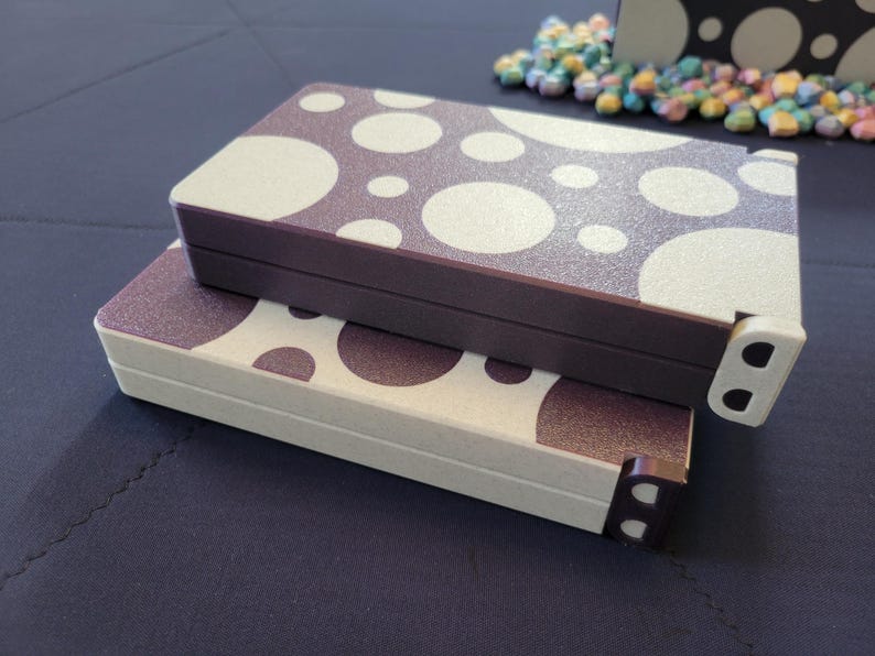 May include: Two rectangular boxes with a polka dot pattern in white and dark brown. The boxes are stacked on top of each other. The edges of the boxes are dark brown and white. The boxes are on a dark blue surface.