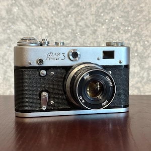 May include: A silver and black FED 3 rangefinder camera featuring a textured black body, a metallic top plate with dials, and a circular lens with the text FED 3, 8591661, and 2,8/52 printed on the front. The camera has a viewfinder and a small lever.
