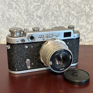 Puede incluir: Una cámara telemétrica vintage FED-2 con una parte superior de metal plateado y un cuerpo negro texturizado. La cámara cuenta con un lente plateado prominente con texto en cirílico, un visor circular y varios diales de ajuste en la placa superior. Una tapa de lente negra se encuentra al lado.