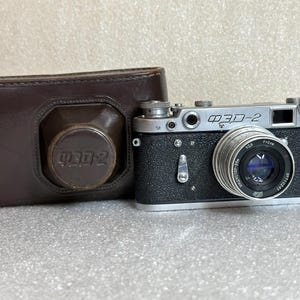 Puede incluir: Una cámara ФЭД-2 vintage con una funda de cuero. La cámara es plateada y negra con una lente y el texto "ФЭД-2". La funda de cuero marrón tiene una tapa de lente a juego con el mismo texto. Es una cámara de película clásica.