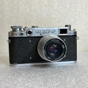 Puede incluir: Una cámara ФЭД-2 vintage en blanco y negro con una lente prominente. El cuerpo de la cámara tiene un acabado texturizado y la lente está rodeada por un anillo plateado. La cámara tiene varios diales y botones.