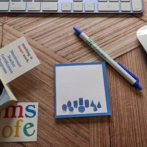 May include: A blue and white notepad with a blue border and a drawing of geometric shapes. The notepad is on a wooden surface with a blue pen and a white keyboard in the background. The text "teaching is heart work" is on a white and red notecard.