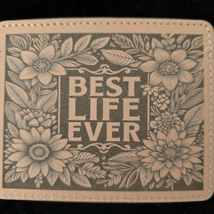 Puede incluir: Un posavasos rectangular de cuero color canela claro con esquinas redondeadas y bordes cosidos. El centro presenta el texto BEST LIFE EVER en una fuente negrita, rodeado por una detallada ilustración grabada de flores y hojas.