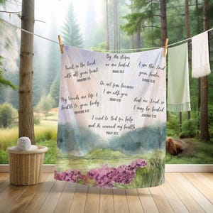 May include: A white and pastel-colored blanket with inspirational Bible verses printed on it. The blanket features a watercolor landscape with purple flowers and a mountain range. The text includes phrases like "Trust in the Lord" and "By His stripes we are healed."