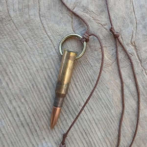 Può includere: Un ciondolo a forma di proiettile in ottone con punta in rame è appeso a una collana con cordino marrone. Il proiettile è attaccato a un anello color oro. La collana è esposta su una superficie di legno.