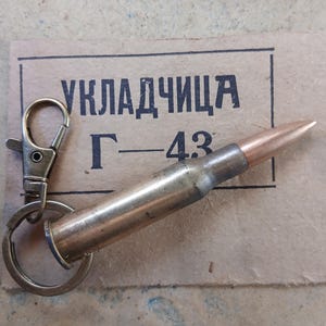 Könnte beinhalten: Ein Schlüsselanhänger mit einer Messingkugel, einem silbernen Karabiner und einem Ring. Die Kugel liegt auf einem Stück beigem Papier mit schwarzem kyrillischem Text und der Nummer "43". Die Kugel hat eine kupferfarbene Spitze.