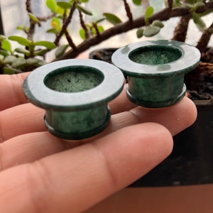 May include: Two dark green stone ear gauges. Each gauge has a flared, cylindrical shape with a flat, circular front and back. The stone has a mottled appearance, with darker and lighter green areas. The gauges are displayed against a blurred background.