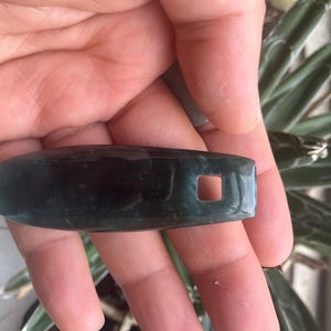 Puede incluir: Un objeto oblongo de color verde oscuro con un agujero cuadrado en un extremo. La superficie pulida refleja la luz, lo que sugiere una piedra preciosa. El objeto se sostiene en una mano. Probablemente sea un llavero.
