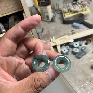 May include: Two green jade ear gauges with a circular design. The gauges are held in a hand, showcasing their polished surface and translucent quality. The background includes workshop tools and materials.