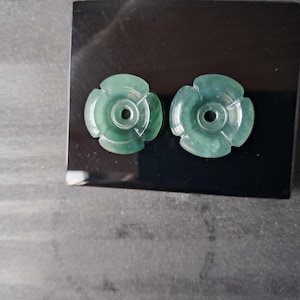 May include: Two circular, translucent green stone pendants, each featuring a carved four-petal flower design with a central circular hole. The pendants exhibit natural color variations and a smooth, polished surface texture throughout.