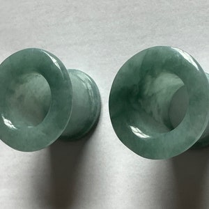 May include: Two jade green ear plugs with a tunnel design. The plugs are made of a translucent stone with natural variations in color. The plugs are smooth and polished, with a rounded shape.