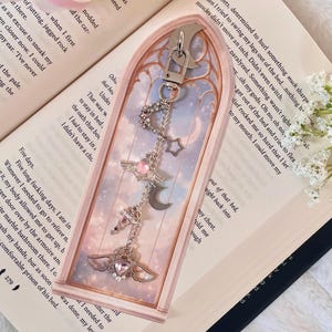 May include: A silver keychain featuring celestial charms, including a crescent moon, stars, and heart-shaped pendants with wings. The keychain is presented in a rectangular, arched display case with a pink and blue backdrop.