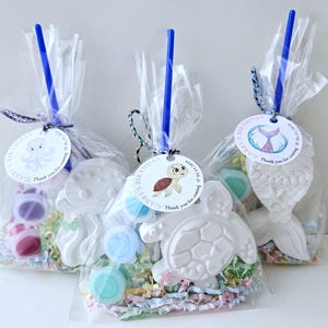 Op de afbeelding: Drie transparante cadeauzakjes vastgebonden met blauw lint en labels. Elke zak bevat een wit zeedier-vormig item, verfpotjes en kleurrijke papiersnippers. De labels tonen illustraties van zeedieren en de tekst "Gabby's Birthday".