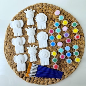 May include: A craft kit featuring white elephant, giraffe, and lion figurines, paint pots in various colors, and paintbrushes. The figurines are arranged on a woven mat, ready for painting and creative fun.