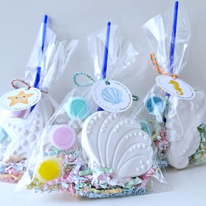 May include: Three clear cellophane bags filled with sea-themed treats. Each bag features a white seashell, starfish, or seahorse, along with colorful candies and shredded paper. Blue sticks and decorative tags complete the party favors.
