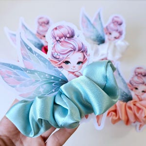 Pode incluir: Uma coleção de scrunchies de cetim em várias cores, incluindo azul claro e pêssego, cada um adornado com um adesivo de fada. As fadas têm cabelo rosa e asas delicadas, adicionando um toque lúdico aos acessórios de cabelo.