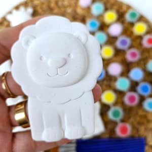 Könnte beinhalten: Ein weißes, löwenförmiges Bastelobjekt mit einem lächelnden Gesicht und einer flauschigen Mähne. Der Löwe wird in einer Hand gehalten, mit bunten Farbpunkten und Pinseln im Hintergrund. Dieser Artikel ist wahrscheinlich für ein DIY-Bastelprojekt.