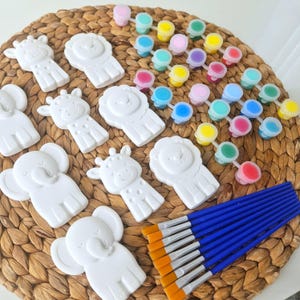 Puede incluir: Colección de figuras blancas con forma de animales, incluyendo elefantes, jirafas y leones, dispuestas sobre una esterilla tejida. También hay pequeños botes de pintura de varios colores y un juego de pinceles con mango azul, lo que sugiere una actividad de manualidades o pintura.