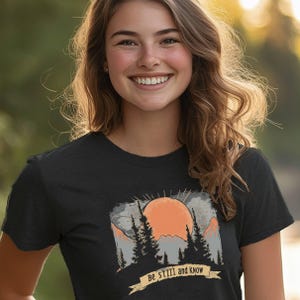 May include: A black t-shirt with a vintage-style graphic of a sunset over mountains and trees. The text "Be still and know" is printed on a banner below the graphic.