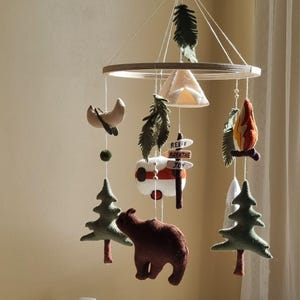 May include: A felt mobile featuring camping-themed elements. Includes a brown bear, a red and white camper, a campfire, a canoe, and trees. The mobile has a wooden ring and is suspended by strings. The sign reads "Relax, Breathe, Joy."