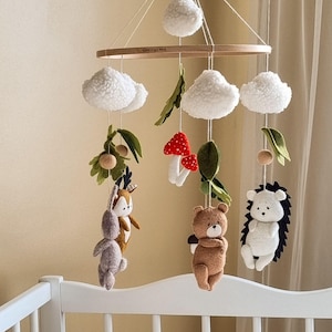 May include: A handmade baby mobile featuring a wooden ring with hanging plush clouds, felt leaves, and woodland creatures. The mobile includes a deer, bear, hedgehog, and mushrooms, all in soft, neutral colors. The mobile is suspended above a white crib.