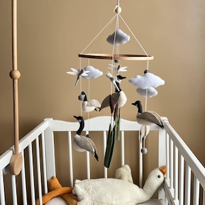 May include: A hanging nursery mobile featuring felt Canada geese, white clouds, dragonflies, and cattails suspended from a circular wooden frame. The mobile includes a wooden arm attachment and hangs above a white crib containing a plush white goose.