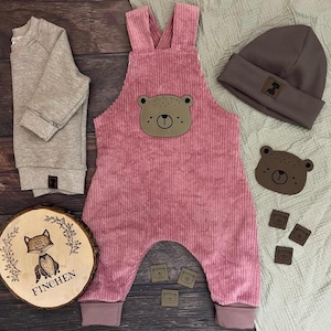 Könnte beinhalten: Eine Kollektion von Babykleidung in Rosa- und Brauntönen. Dazu gehören eine rosa Cord-Latzhose mit Bärenkopf, ein graues Sweatshirt, eine braune Mütze und ein Holzschild mit dem Wort "FINCHEN".