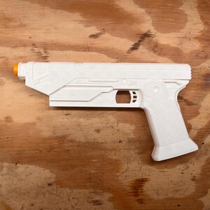 May include: A white plastic toy blaster with a rectangular barrel and a textured grip. The toy features a small orange tip at the front of the barrel and a hollow trigger guard area. The design includes molded geometric lines and circular details.