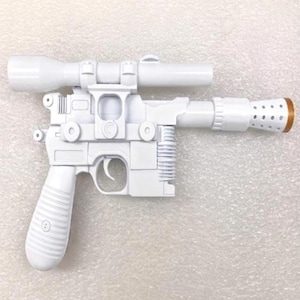 May include: A white toy blaster with a long top-mounted scope, a textured grip, and a perforated barrel tip featuring a brown circular accent. The plastic construction includes a trigger, a side-mounted cooling vent detail, and a blocky frame.