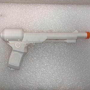 Peut inclure: Un pistolet jouet blanc doté d'un long canon et d'une poignée texturée. Le pistolet présente une section arrière cylindrique et un petit embout orange vif à l'extrémité du canon. Le design est épuré et monochrome, à l'exception de l'embout orange.