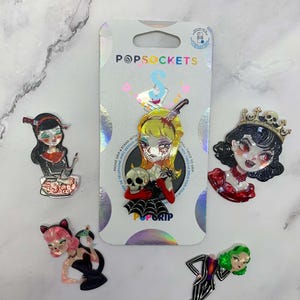 May include: A collection of five character-themed phone grips, including one attached to its original PopSockets packaging. The designs feature stylized figures with various motifs, including skulls, spiders, crowns, and knives, rendered in vibrant colors.