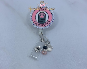 May include: A retractable badge reel with a pink and white circular design. The design features a gray paw print and the word "PAWSITIVE". The reel is embellished with rhinestones and charms, including a paw print and beads.