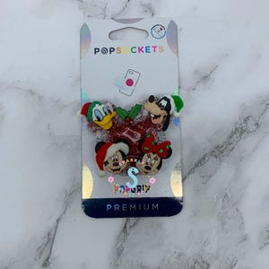 May include: A PopSockets PopGrip phone accessory featuring a red glittery Mickey Mouse head shape adorned with holiday-themed Disney characters including Donald Duck, Goofy, Mickey Mouse, and Minnie Mouse, all wearing festive Christmas hats.