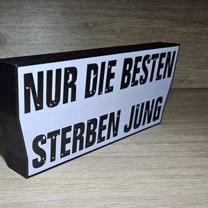 Könnte beinhalten: Ein schwarz-weißes Schild mit dem deutschen Text "NUR DIE BESTEN STERBEN JUNG". Das Schild hat einen weißen Hintergrund mit schwarzem Text und einen schwarzen Rand. Das Schild ist abgewinkelt.
